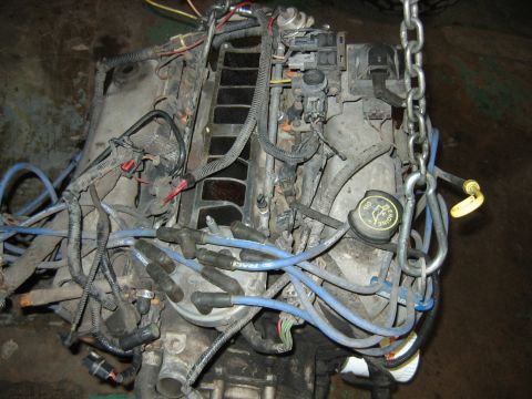 1992 Ford Bronco - A removed engine block sits on a shop floor, supported by a lifting chain, showing blue spark plug wires labeled 'RACING', a black oil fill…