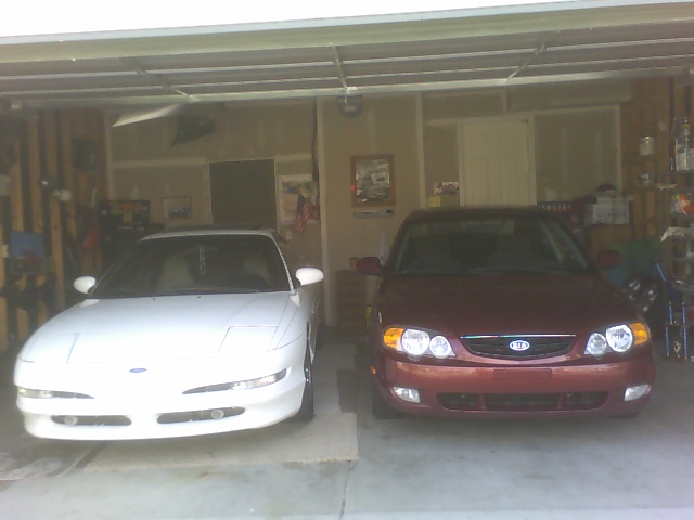 White car and red car parked in a garage. The red car has a Kia logo