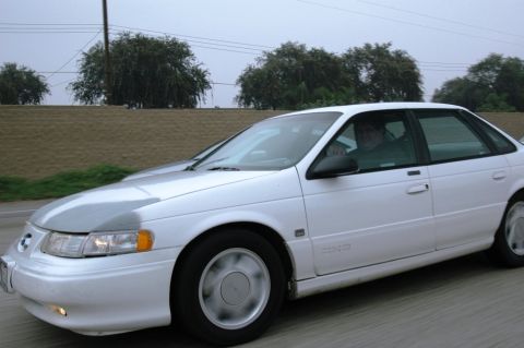 1991 Ford Taurus - White 1991 Ford Taurus sedan captured in motion on a highway during daytime.