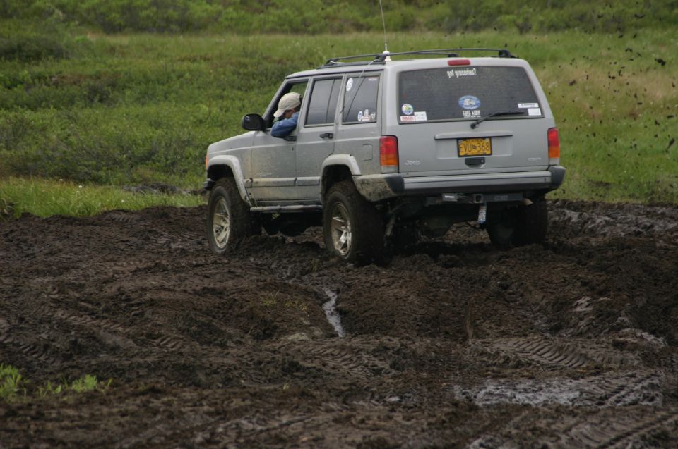 Silver Jeep Cherokee XJ with lifted suspension driving through deep mud. License plate reads EVU-368