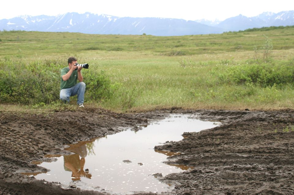 Man with telephoto camera crouching beside a muddy puddle in a green tundra valley