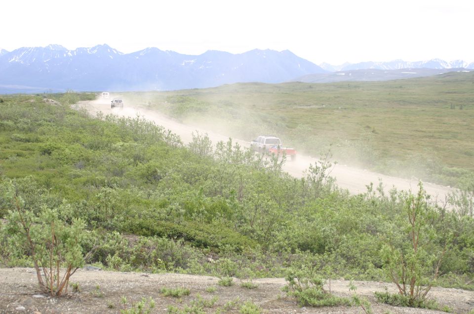 Two vehicles drive on a dusty dirt road with mountains in the background