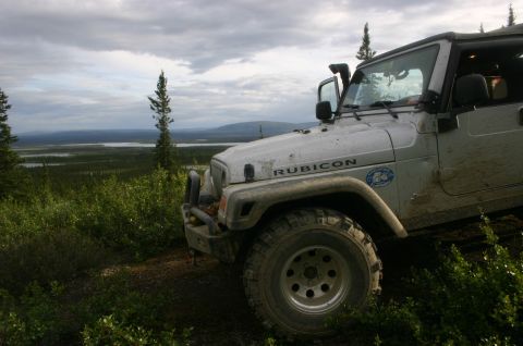 2003 Jeep TJ - A silver Jeep Wrangler with a Rubicon decal is parked on a grassy hill, overlooking a vast landscape of trees and a lake under a cloudy sky.