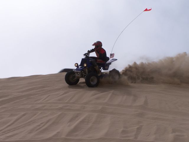 Rider on quad bike kicking up dust on a sand dune with an American flag and a red flag