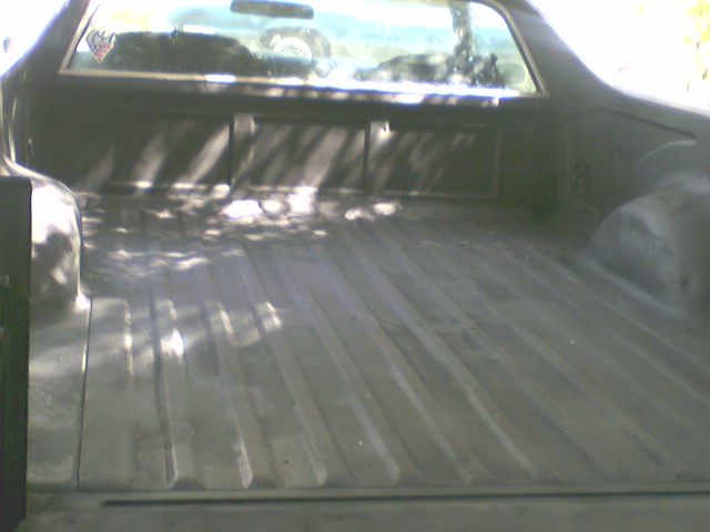 Empty ribbed cargo bed of a Ford Ranchero viewed from the rear, with dappled shade and cab rear window visible.