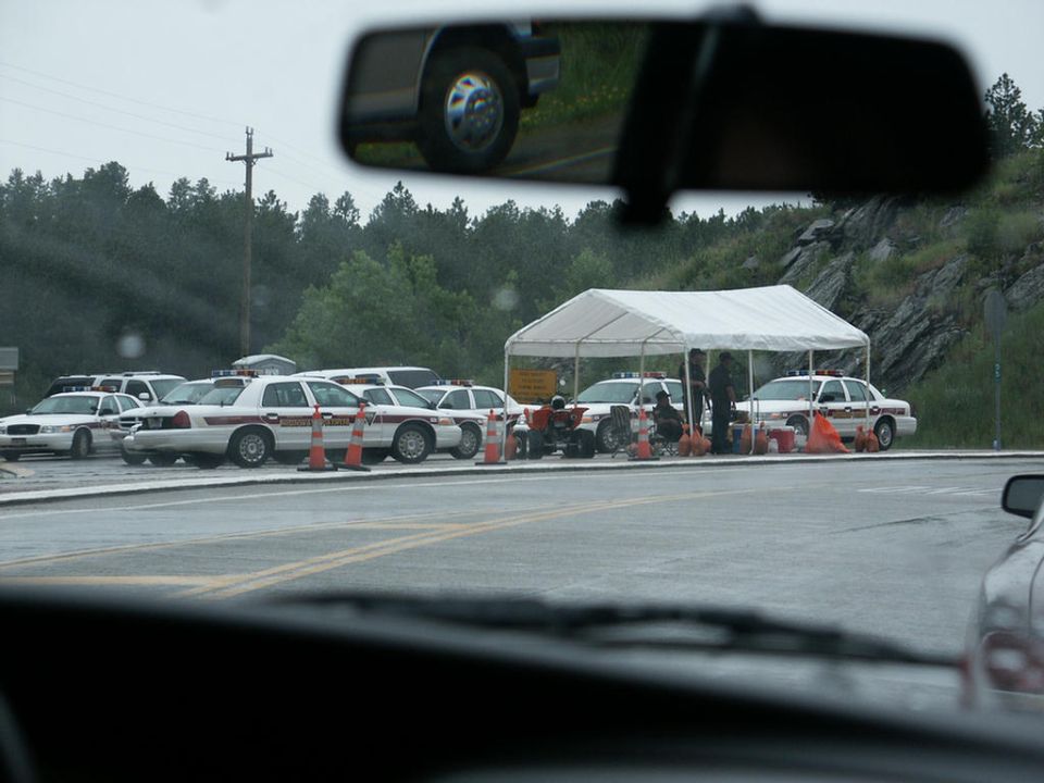 Several police cars are parked in a lot under a white tent, with orange cones around them