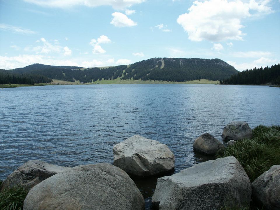 Lake with rocks in foreground and forested mountains under a cloudy sky