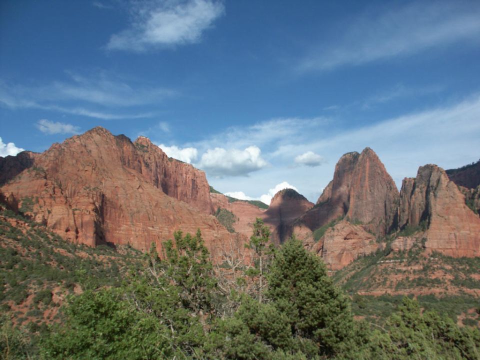 Red rock mountains under a blue sky with scattered clouds, foreground of green trees