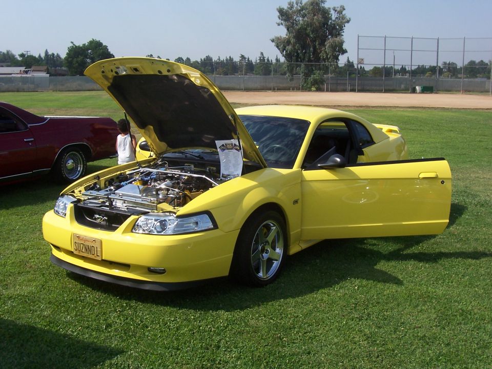 Yellow Ford Mustang with hood and driver's door open, showing engine and interior. License plate reads SUZNNOL