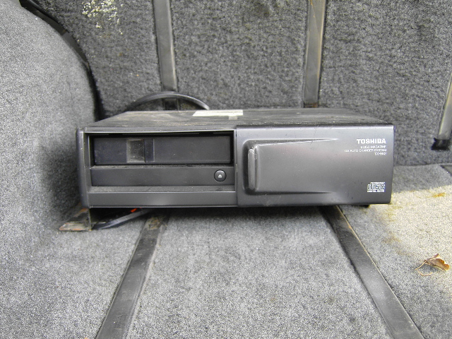 Toshiba CD changer mounted on carpeted floor with straps. Text reads TOSHIBA CD AUTO CHANGER SYSTEM TC-460