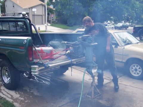 1972 Ford Ranchero - Man tests outboard motor in the bed of a green pickup truck, with a tan car parked nearby.