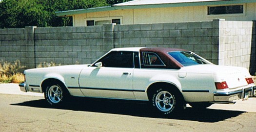 White two-door coupe with maroon vinyl roof, side view, parked by a block wall