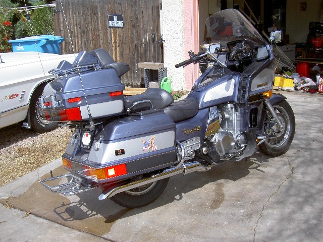 Gray Kawasaki Voyager touring motorcycle from rear three-quarter angle in driveway, white classic car partially visible at left