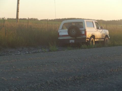1990 Ford Bronco - A white 1990 Ford Bronco is parked off a dirt road, with a spare tire mounted on the rear.