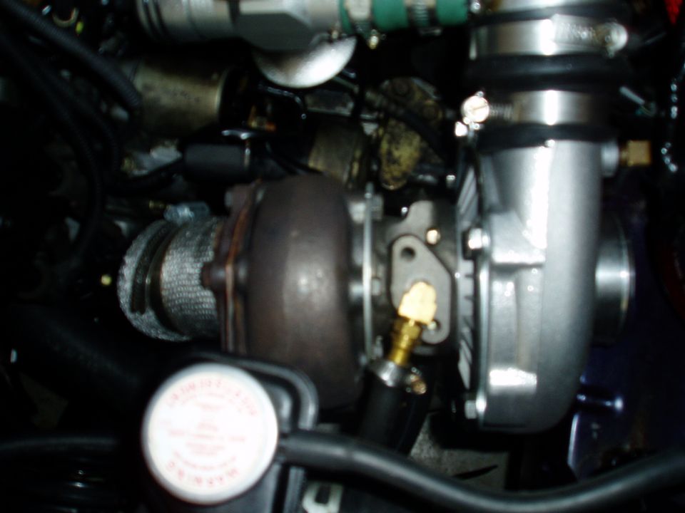 Turbocharger with heat wrap and silver housing, connected to engine components. A white cap with red text is visible