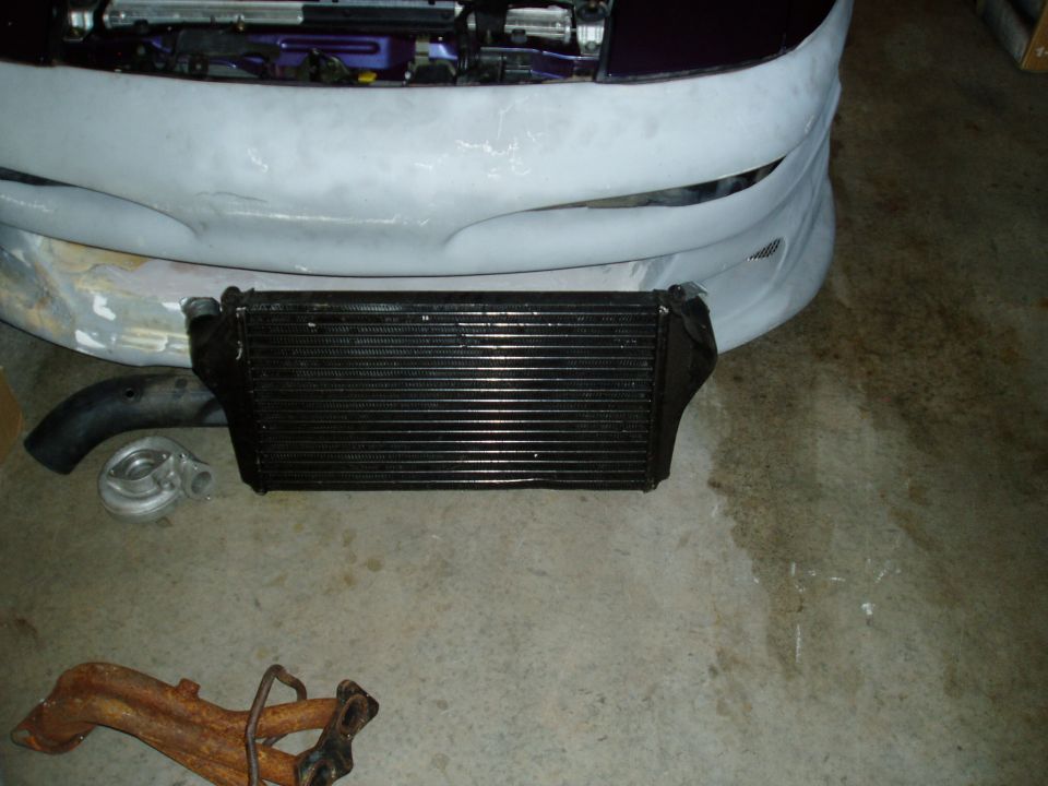 Intercooler, turbocharger, and exhaust manifold parts laid out in front of a car's bumper