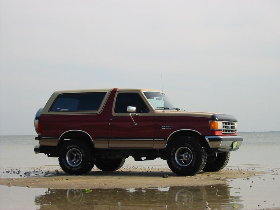 Two-tone red and tan Ford Bronco SUV parked on a wet beach, side view with white-letter tires.