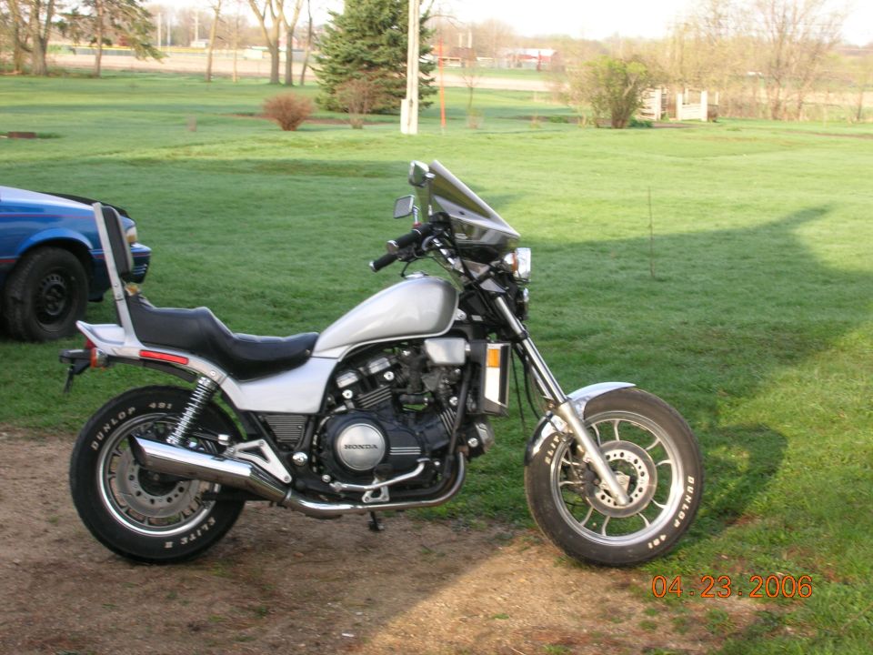 Silver motorcycle with a black seat and windshield, parked on dirt next to a blue car