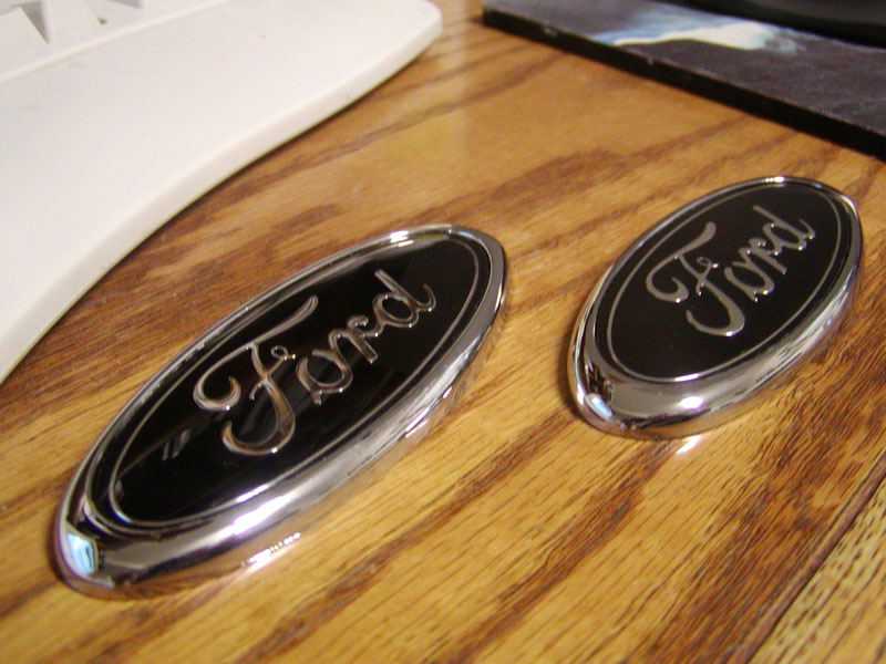 Two chrome Ford oval emblems with black backgrounds sitting on a wood surface