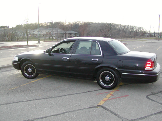 Black Ford Crown Victoria sedan in a parking lot, side profile with steel wheels and bare trees behind