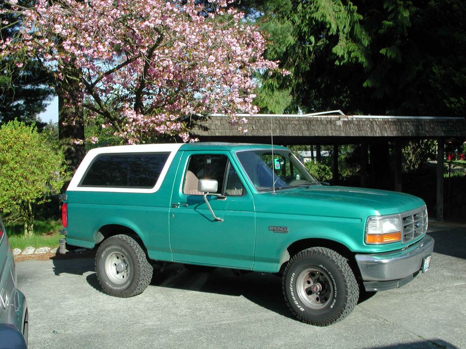 Teal Ford Bronco with white top parked on driveway, passenger side view, pink blossom tree behind.