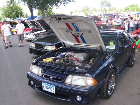 1989 Ford Mustang - A dark blue Fox-body Ford Mustang GT hatchback sits hood-up at a car show, showing a 5.0 with an aftermarket intake, blue silicone hose and…