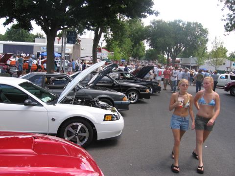 1989 Ford Mustang - A row of Mustangs parked hood-up along a tree-lined street at a summer car show, led by a white New Edge coupe on machined five-spoke wheel…