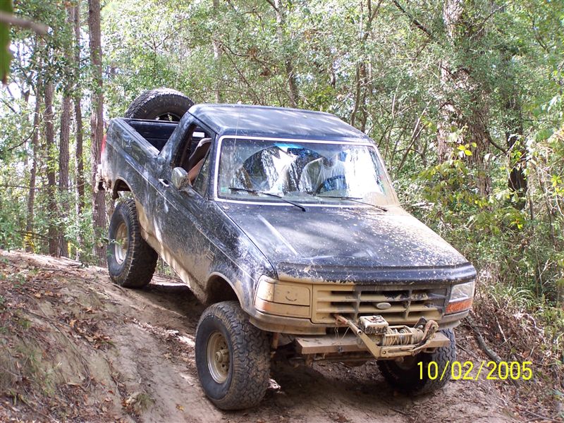 Muddy topless dark green Ford Bronco with front winch climbing a dirt bank on a trail