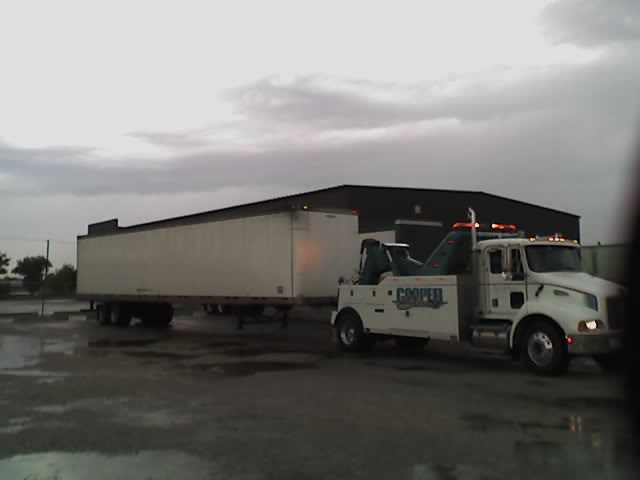White tow truck with 'COOPER' text on the side, towing a white trailer. The ground is wet