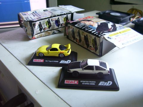 1994 Ford Probe - Two collectible toy cars, a yellow Mazda RX-7 and a white Toyota Sprinter Trueno, are displayed with their original anime-themed packaging.