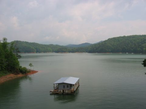 1985 Ford Crown Victoria LTD - A houseboat sits on a large lake with rolling, forested hills in the background under an overcast sky.