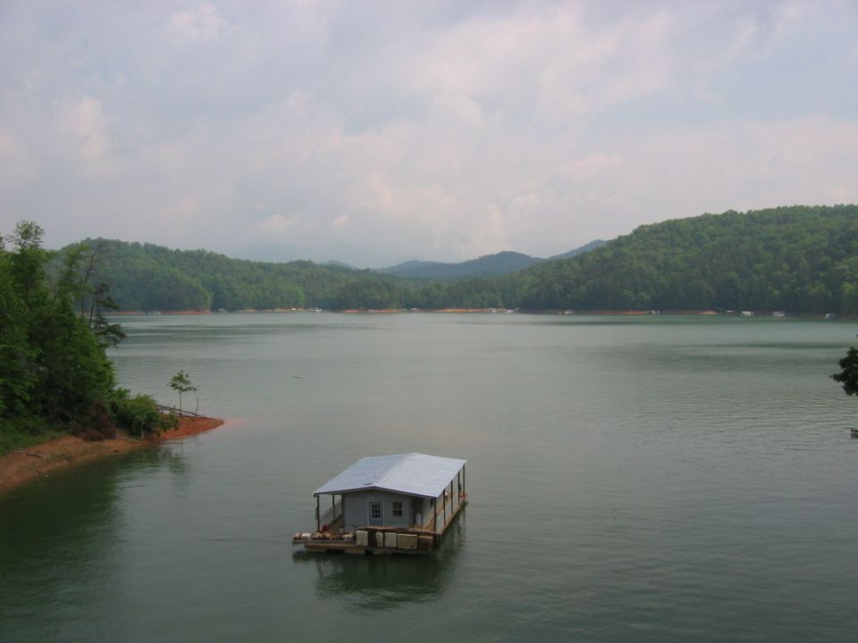 Houseboat with a metal roof floats on a calm lake, surrounded by tree-covered hills under a cloudy sky