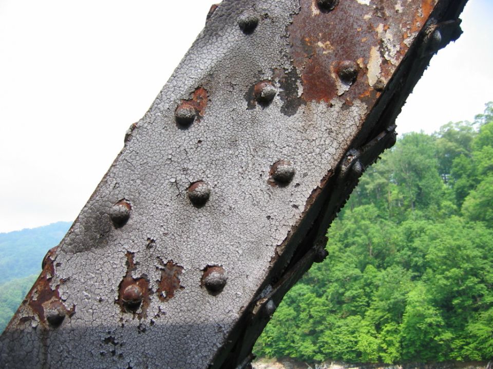 Close-up of a heavily rusted metal beam with peeling paint and rivets. Lush green trees and a body of water are visible in the background
