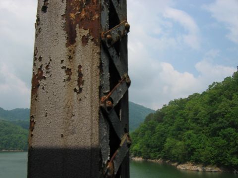 1985 Ford Crown Victoria LTD - A close-up of a weathered metal support structure overlooks a lake and tree-covered mountains under a cloudy sky.
