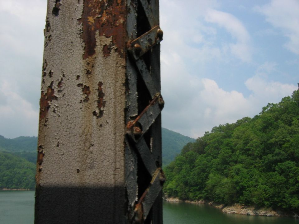 Rusted metal structure with peeling paint against a backdrop of green hills and a lake