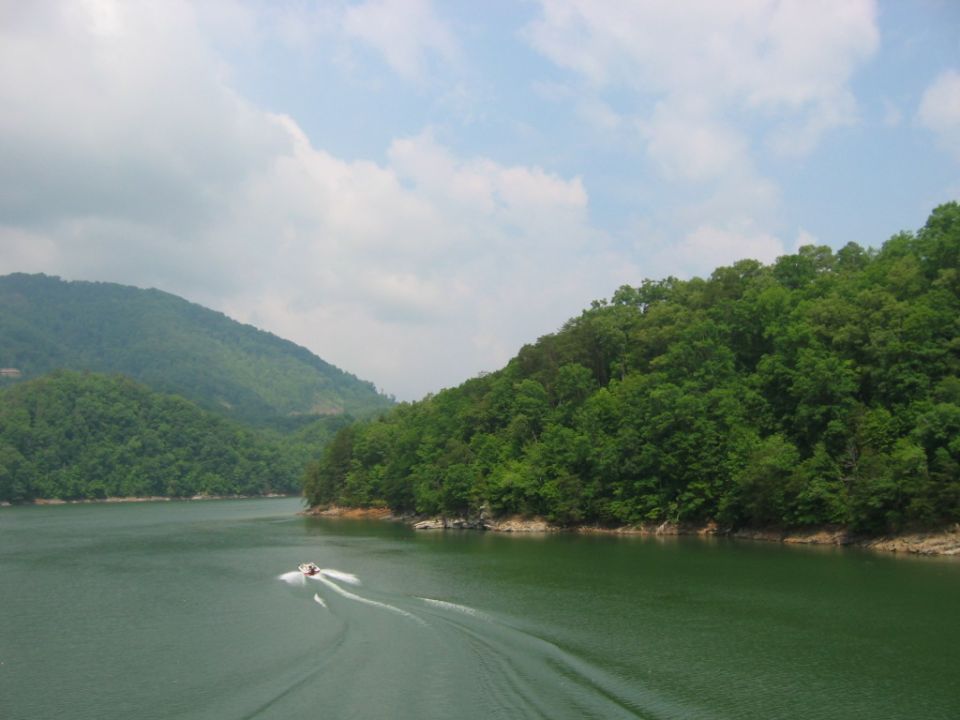 Boat speeds across a green lake, leaving a wake, with forested hills under a cloudy sky