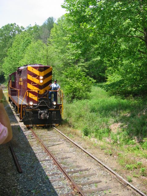 1985 Ford Crown Victoria LTD - A train with a worker on the front travels down the tracks through a wooded area.