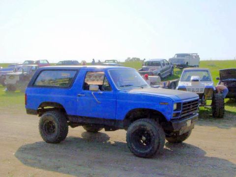 1983 Ford Bronco - A blue SUV sits in a dusty lot surrounded by other vehicles under a bright sky.