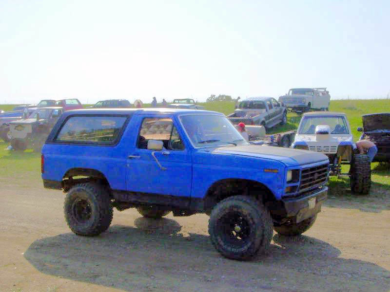 Blue SUV with aftermarket wheels and tires, parked in a lot with other vehicles. Some people are working on a vehicle in the background