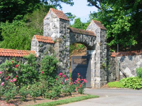 1985 Ford Crown Victoria LTD - A stone gatehouse entrance with a tiled roof, surrounded by lush vegetation and blooming roses.