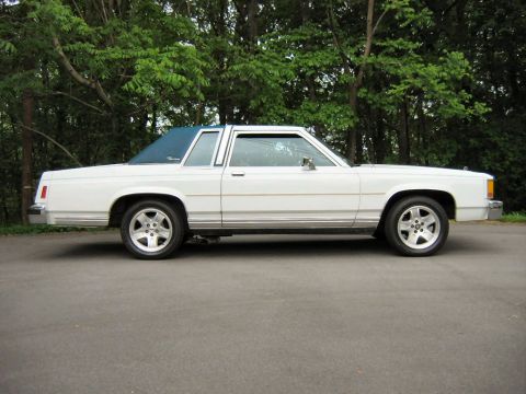 1985 Ford LTD Crown Victoria - A white 1983 Ford sedan with a blue vinyl roof is parked on an asphalt surface, with a backdrop of lush green trees.