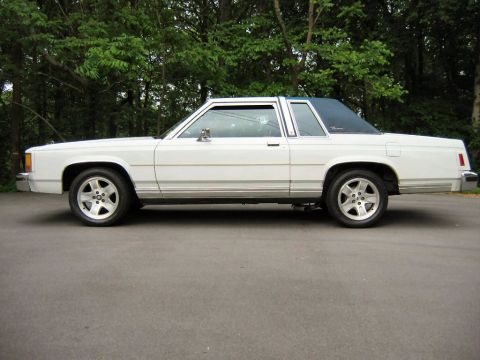 1985 Ford LTD Crown Victoria - White two-door Ford full-size coupe shot in full profile from the driver's side on asphalt in front of dense green trees. It has a dark pad…
