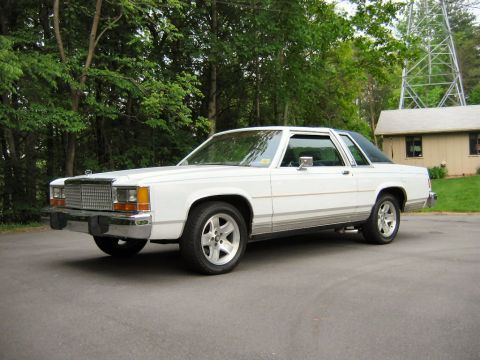 1985 Ford LTD Crown Victoria - A white two-door sedan with a dark roof is parked on an asphalt surface, with trees and a building in the background.