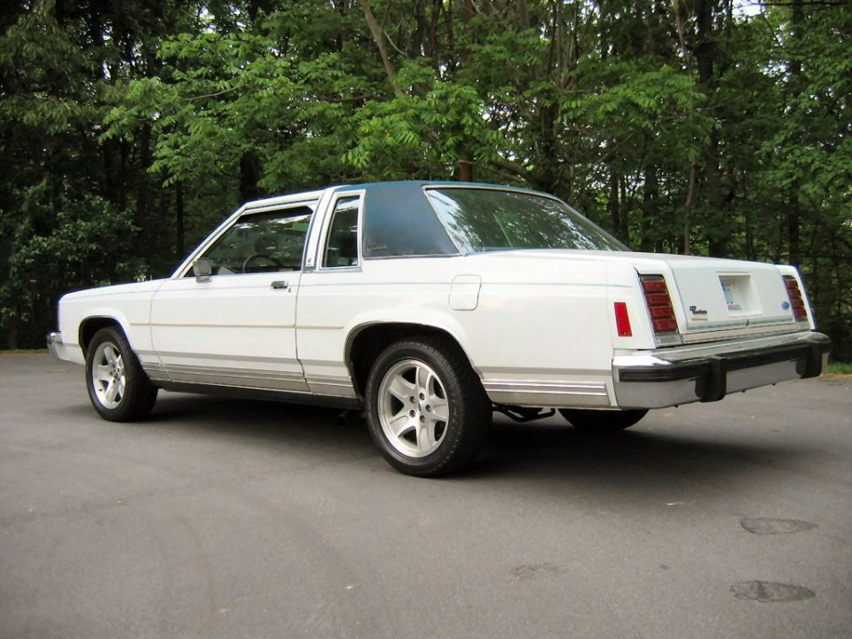 White 1985 Ford Crown Victoria LTD with a blue roof, parked on asphalt with trees in the background