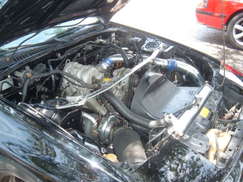 1989 Ford Probe - The engine bay of a black car is shown, a turbocharger and intercooler piping, with a red car visible in the background.