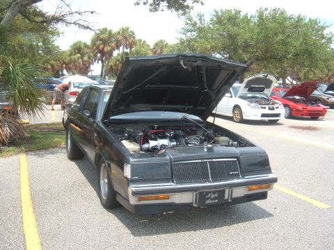 1989 Ford Probe - A black Buick Regal Grand National is parked with its hood open, showing the engine. Other cars are visible in the background.