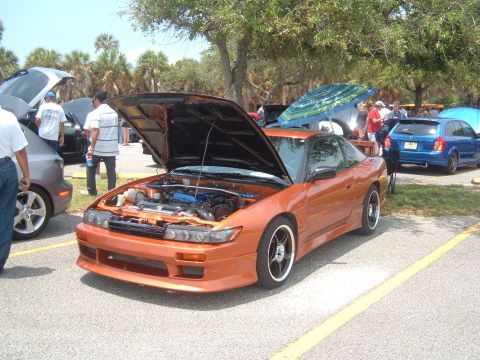 1989 Ford Probe - An orange sports coupe is parked with its hood open, revealing the engine, at an outdoor car gathering.