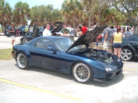 1989 Ford Probe - A dark blue Mazda RX-7 is displayed with its hood open at an outdoor car event, surrounded by spectators.