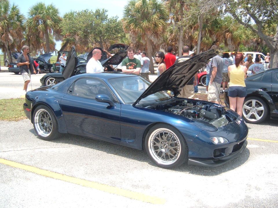 Blue Mazda RX-7 with hood open, showing engine. People gathered around at an outdoor car show