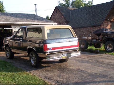 1989 Ford Bronco - A dark green and tan Ford Bronco is parked in a driveway with a second vehicle chassis visible in the background.